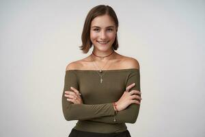 Indoor photo of young attractive brown haired female keeping her hands crossed while posing over white background, looking positively at camera with charming smile