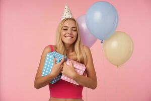 Disparo de Encantado bonito largo peludo mujer con casual peinado celebrando cumpleaños con multicolor aire globos, demostración contento reacción en consiguiendo increíble regalos, aislado terminado rosado antecedentes foto