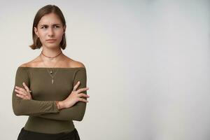 Bewildered young dark haired lady with short haircut folding hands on her chest while looking confusedly aside, dressed in olive blouse while posing over white background photo