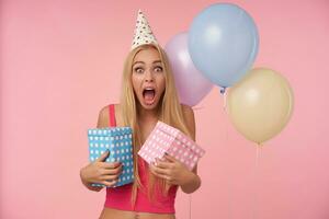 Studio photo of overjoyed young blonde female in pink top and holday cone cap rejoicing while posing in multicolored air balloons, looking at camera happily with wide eyes and mouth opened