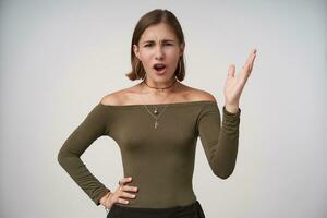 Irritated young dark haired lady with short haircut frowning her eyebrows while looking at camera and raising emotionally her palm, isolated over white background photo