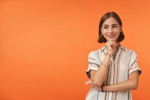 linda mujer curioso sonrisa vistiendo a rayas camisa, anillos y esposas plegable manos y conmovedor su mentón. sonriente niña mirando a el izquierda sitio para tu comercial terminado naranja antecedentes foto