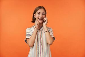 Female student talk on smart phone, wearing striped shirt, teeth braces and bracelets, hear a good news. Smiling girl looking happy and watching at the camera isolated over orange background photo
