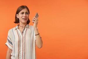 Unhappy girl hold and look to the smartphone. Did not like what she hear and shows it. Wearing striped shirt, teeth braces and bracelets. Watching to the right, copy space over orange background photo