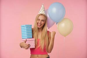Cheerful pretty young blonde woman rejoicing nice party together with friends, being in high spirit and raising fist happily, isolated over pink background and bunch of multicolored helium balloons photo