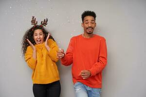 Indoor photo of joyful young dark skinned curly pair making confetti with clapper while celebrating new year, wearing cosy woolen colourful sweaters and holiday hoop over grey background