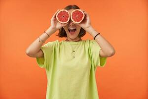 Female student, young lady with short brunette hair holding grapefruit over her eyes. Surprised looking. Standing over orange background. Wearing green t-shirt, teeth braces and bracelets photo