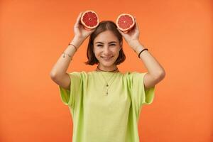 Teenage girl, cheerful and happy with short brunette hair holding grapefruit over her head. Standing over orange background. Wearing green t-shirt, teeth braces and bracelets photo