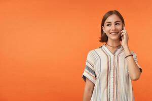 atractivo joven mujer hablar en un teléfono inteligente y mirando a el izquierda a el Copiar espacio terminado naranja antecedentes. teniendo un bonito hablar. vistiendo a rayas camisa, dientes tirantes y esposas. foto