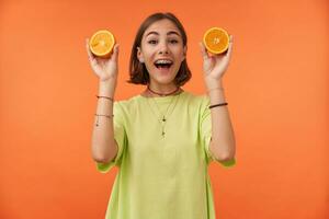 Portrait of attractive, nice looking girl with short brunette hair showing oranges and smile. Getting ready to make some juice. Wearing green t-shirt, teeth braces and bracelets photo