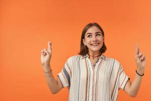 Young pretty woman with short hair crossing fingers and smile looking to the left upper corner at the copy space over orange back ground. Wearing striped shirt, teeth braces and bracelets photo