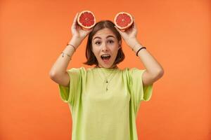 Portrait of attractive, shocked and funny, nice looking girl with short brunette hair. Holding grapefruit over her head. Standing over orange background. Wearing green t-shirt, braces and bracelets photo