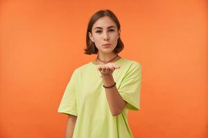 Female student, young lady with short brunette hair watching at the camera isolated over orange background. Sending kiss, showing interest. Wearing green t-shirt, necklace, bracelets and rings photo