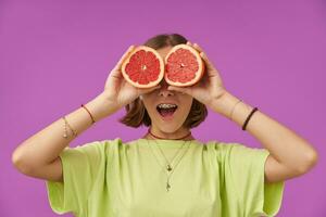 Teenage girl, cheerful and happy, woman with brunette short hair holding grapefruit over her eyes. Standing over purple background. Wearing green t-shirt, teeth braces, bracelets and necklace photo