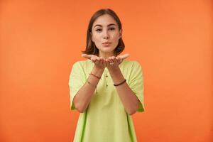 Portrait of attractive, nice looking girl with short brunette hair, sending a kiss, tempting. Look to the camera over orange background. Wearing green t-shirt, bracelets and rings photo