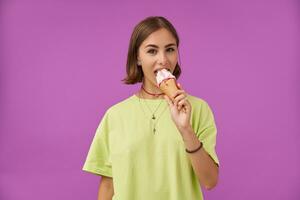 Teenage girl, cheerful and happy, with brunette short hair. Eat an ice cream and looking to the camera, over purple background. Wearing green t-shirt, bracelets, rings and necklace photo