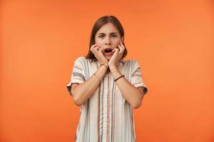 Young pretty woman with short hair touch her face with both hands, open her mouth. Hear something exciting. Wearing striped shirt and bracelets. Watching at the camera isolated over orange background photo