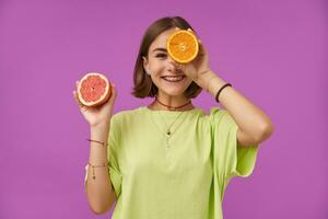 Portrait of attractive, cute girl with short brunette hair. Holding orange over her eye, cover one eye. Standing over purple background. Wearing green t-shirt, necklace, braces and bracelets photo