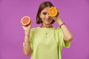 Portrait of attractive, cute girl with short brunette hair. Holding orange over her eye, cover one eye. Standing over purple background. Wearing green t-shirt, necklace, braces and bracelets photo