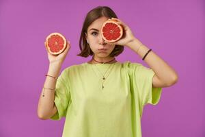 Female student, young lady with short brunette hair. Holding grapefruit over her eye, looking sad. Standing over purple background. Wearing green t-shirt, necklace, braces and bracelets photo