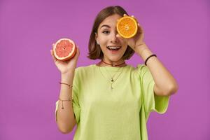Female student, young surprised lady with short brunette hair. Holding orange over her eye, cover one eye. Standing over purple background. Wearing green t-shirt, necklace, braces and bracelets photo