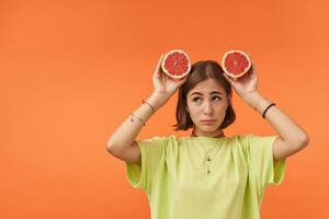 Young pretty woman with short brunette hair, looks sad, upset. Holding grapefruit over her head. Standing over orange background. Wearing green t-shirt, braces and bracelets photo