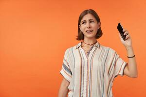 Adult girl looking unhappy and hanging up a phone. Unlike call. Student pick up a phone, wearing striped shirt, teeth braces and bracelets. Watching to the left at copy space over orange background photo