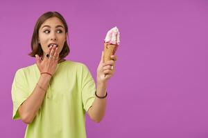 Teenage girl, surprised lady, with brunette short hair, holding an ice cream and looking at the right to the copy space over purple background. Wearing green t-shirt, bracelets, rings and necklace photo