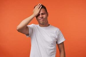 Stressed young short haired brunette guy keeping his eyes closed while frowning face and holding palm on forehead, standing isolated over orange background photo