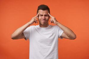 Unhappy young handsome brown haired man with short haircut keeping fingers on temples and frowning face while standing over orange background in white basic t-shirt photo