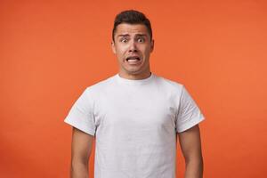 Frightened young brown-eyed brunette man with short haircut rounding amazedly his eyes while looking scaredly at camera, standing over orange background photo