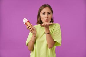 Portrait of attractive, nice looking girl with ice cream. Sanding a kiss to the camera and standing over purple background. Wearing green t-shirt, bracelets, rings and necklace photo