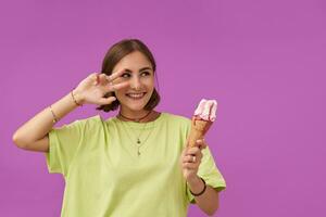 Teenage girl, happy, with brunette hair. Lady with ice cream showing a signal, watching to the right at the copy space over purple background. Wearing green t-shirt, bracelets, rings and necklaces photo