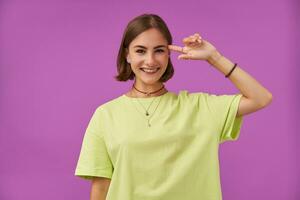 Adolescente chica, alegre y feliz, con morena corto cabello. participación un dedo siguiente a su templo. en pie terminado púrpura antecedentes. vistiendo verde camiseta, dientes tirantes, collar, esposas y anillos foto