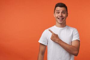 Horizontal shot of young brown-eyed short haired guy showing aside with index finger while looking joyfully at camera, standing over orange background photo