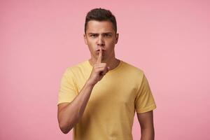 Severe young handsome short haired brunette man raising hand with hush gesture to his mouth and frowning eyebrows while looking seriously at camera, isolated over pink background photo