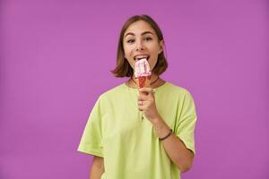 Portrait of attractive, nice looking girl. Looking to the camera and licking an ice cream, over purple background. Wearing green t-shirt, teeth braces, bracelets, rings and necklace photo