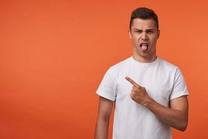 Displeased young short haired brunette man frowning his face and sticking out tongue while pointing aside with forefinger, isolated over orange background photo