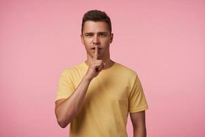 Indoor photo of young brunette guy dressed in casual wear keeping raised index finger on his mout while looking at camera, isolated over pink background