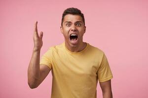 Irritated young short haired brunette man with short haircut screaming crossly with wide mouth opened and raising emotionally palm while posing over pink background photo