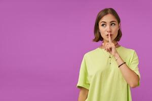 Portrait of attractive, nice looking girl. Touching lips with finger, showing a silence sign. Watching to the left at the copy space over purple background. Wearing green t-shirt, bracelets and rings photo