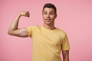 Self-confident young pretty short haired brunette guy keeping hand raised while showing his power and looking glaldy at camera, isolated over pink background photo