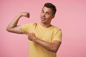 Cheerful young brown-eyed brunette male with short haircut smiling happily at camera while pointing with forefinger on his strong biceps, isolated over pink background photo