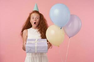 Studio shot of redhead female kid with long hair wearing festive clothes and birthday cap, celebrates holiday, isolated over pink backgroundlooking with air balloons, looking at camera joyfully photo