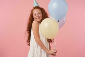 Cheerful curly redhead female kid with long hair holding colored air balloons while standing over pink background, looking in camera happily, expresses true positive emotions photo