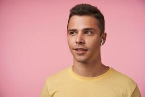 Portrait of positive young handsome short haired brunette male with earpiece looking wonderingly aside while standing over pink background with hands down photo