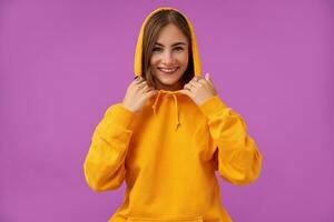 retrato de atractivo, bonito mirando niña con corto morena cabello. sonriente a él cámara y conmovedor capucha con manos, terminado púrpura antecedentes. vistiendo naranja sudadera, anillos y dientes tirantes foto