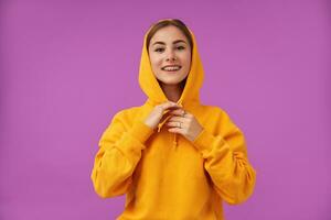 Teenage girl, cheerful and happy, with brunette short hair. Smiling at he camera and touching hood with hands, over purple background. Wearing orange hoodie, rings and teeth braces photo