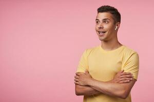 Studio shot of young brunette male with headphones looking emotionally ahead with wide smile and keeping hands folded on chest, isolated over pink background photo