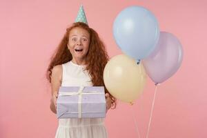 Portrait of joyful little girl with long foxy hair holding gift-wrapped box being excited and surprised to get birthday present, isolated over pink background in white elegant dress photo